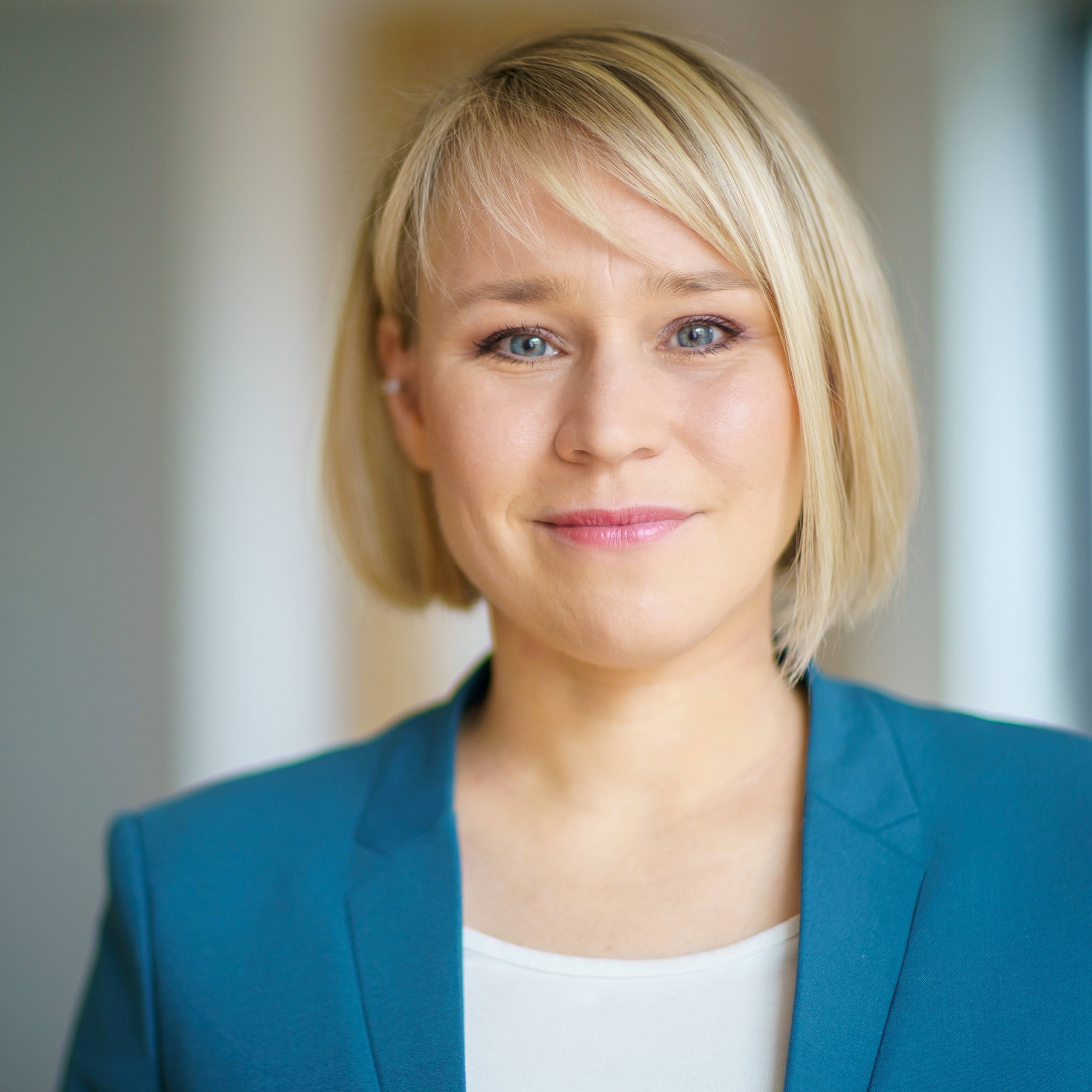 Eine Frau mit kurzen blonden Haaren lächelt in die Kamera. Sie trägt einen blaugrünen Blazer über einem weißen Oberteil. Der Hintergrund ist sanft verschwommen, was dem Porträt ein professionelles, sauberes Aussehen verleiht.