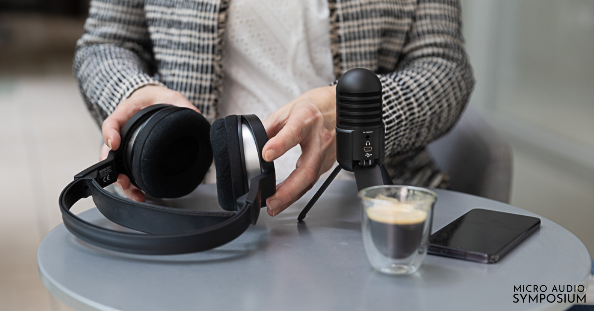 Person in weißem Hemd, die ein Paar schwarze Kopfhörer mit schwarzem Mikrofon, ein Smartphone und eine Tasse Kaffee auf einem runden Tisch hält. Im Hintergrund ist ein Logo mit dem Text „MICRO AUDIO SYMPOSIUM“ zu sehen.