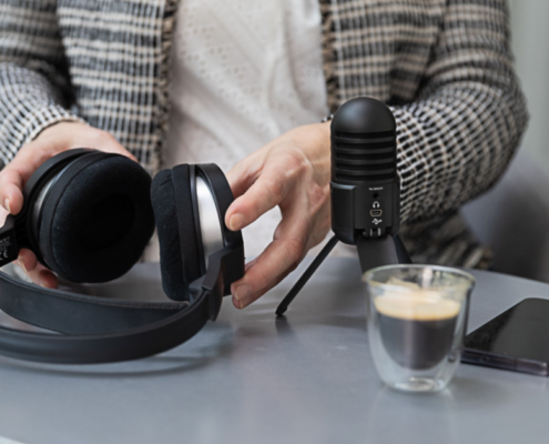 Person in weißem Hemd, die ein Paar schwarze Kopfhörer mit schwarzem Mikrofon, ein Smartphone und eine Tasse Kaffee auf einem runden Tisch hält. Im Hintergrund ist ein Logo mit dem Text „MICRO AUDIO SYMPOSIUM“ zu sehen.