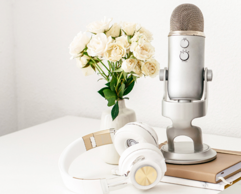 Ein silbernes Mikrofon auf einem Ständer steht auf einem weißen Schreibtisch neben einer Vase mit weißen Rosen und verkörpert die Essenz von Audio im Geschäftsleben. In der Nähe ruht ein Paar weiße Kopfhörer auf zwei gestapelten Notizbüchern. Auf der rechten Seite ist teilweise ein Laptop mit offenem Bildschirm sichtbar.