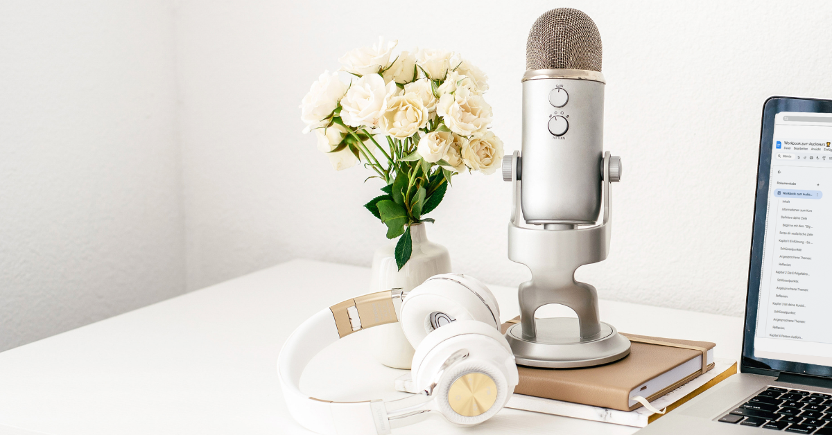 Ein silbernes Mikrofon auf einem Ständer steht auf einem weißen Schreibtisch neben einer Vase mit weißen Rosen und verkörpert die Essenz von Audio im Geschäftsleben. In der Nähe ruht ein Paar weiße Kopfhörer auf zwei gestapelten Notizbüchern. Auf der rechten Seite ist teilweise ein Laptop mit offenem Bildschirm sichtbar.