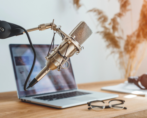 Ein professionelles Mikrofon an einem Galgenarm steht neben einem offenen Laptop auf einem Schreibtisch, daneben stehen Kopfhörer und eine Brille – perfekt für ein Coaching-Geschäfts-Setup. Im Hintergrund sind getrocknete Pflanzen zu sehen, durch die Sonnenlicht strömt, was eine warme Atmosphäre schafft. Der Text „Micro Audio Symposium“ ist sichtbar.