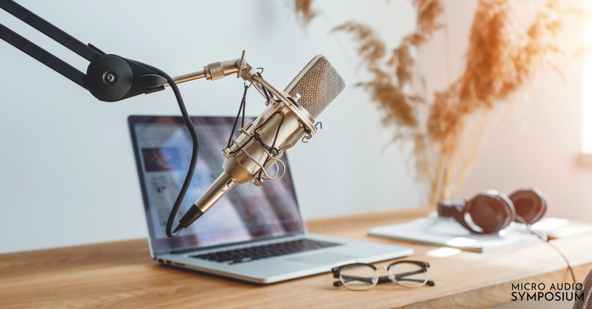 Ein professionelles Mikrofon an einem Galgenarm steht neben einem offenen Laptop auf einem Schreibtisch, daneben stehen Kopfhörer und eine Brille – perfekt für ein Coaching-Geschäfts-Setup. Im Hintergrund sind getrocknete Pflanzen zu sehen, durch die Sonnenlicht strömt, was eine warme Atmosphäre schafft. Der Text „Micro Audio Symposium“ ist sichtbar.