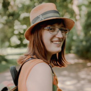 Eine Frau mit braunen Haaren, braunem Hut, Brille und orangefarbenem Oberteil lächelt, während sie im Freien auf einem sonnenbeschienenen Weg mit Grün im Hintergrund steht und diskret Micro Audio-Kopfhörer trägt.