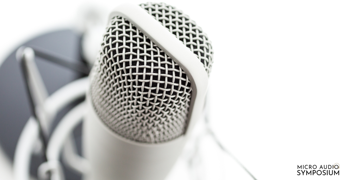 Nahaufnahme eines silbernen Mikrofons mit Gitter und weißer Stoßdämpferhalterung vor unscharfem Hintergrund. „MICRO AUDIO SYMPOSIUM“ und „Micro-Content“ werden in der unteren rechten Ecke angezeigt.
