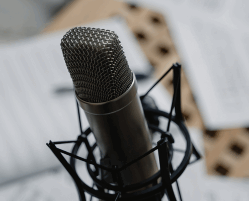 Nahaufnahme eines professionellen Mikrofons auf einem Ständer, das auf eine Podcast-Aufnahme hindeutet, mit unscharfen Papierbögen im Hintergrund und dem Text "MICRO AUDIO SYMPOSIUM" in der unteren rechten Ecke.