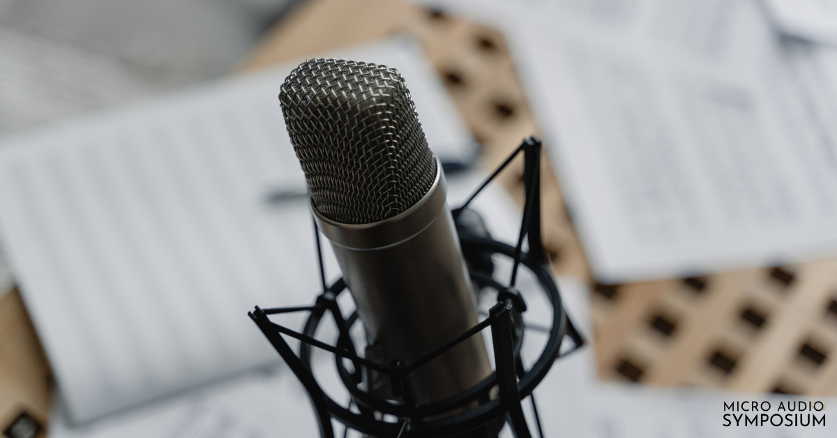 Nahaufnahme eines professionellen Mikrofons auf einem Ständer, das auf eine Podcast-Aufnahme hindeutet, mit unscharfen Papierbögen im Hintergrund und dem Text "MICRO AUDIO SYMPOSIUM" in der unteren rechten Ecke.