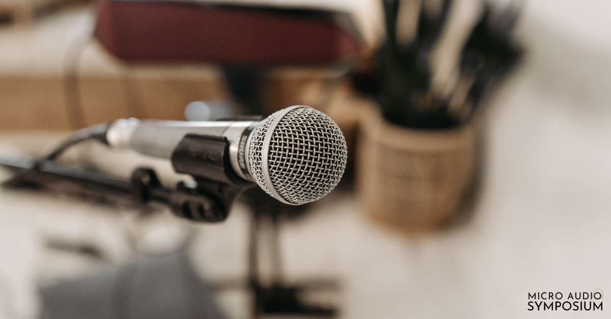 Eine Nahaufnahme eines Mikrofons auf einem Ständer mit einem unscharfen Hintergrund von Gegenständen in Innenräumen. In der unteren rechten Ecke erscheint der Schriftzug "MICRO AUDIO SYMPOSIUM" - die perfekte Inspiration für alle, die Audiokurse erstellen möchten.