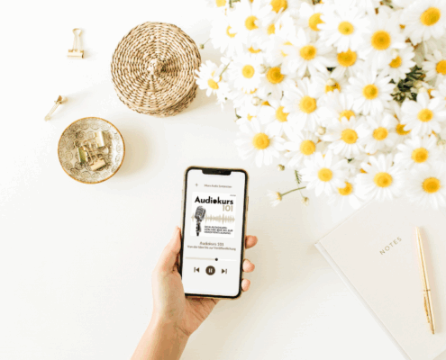 Eine Hand hält ein Smartphone mit einer Hörbuch-App auf einem weißen Schreibtisch mit Gänseblümchen, einem Notizbuch mit einem Stift, Büroklammern in einer Schale und einem geflochtenen Korb. In der rechten unteren Ecke steht "MICRO AUDIO SYMPOSIUM".
