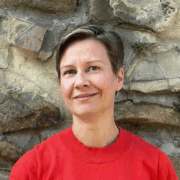 Eine Frau mit kurzen braunen Haaren und rotem Pullover steht mit einem sanften Lächeln vor einer Steinmauer und hält ein diskretes Micro Audio Gerät in der Hand.