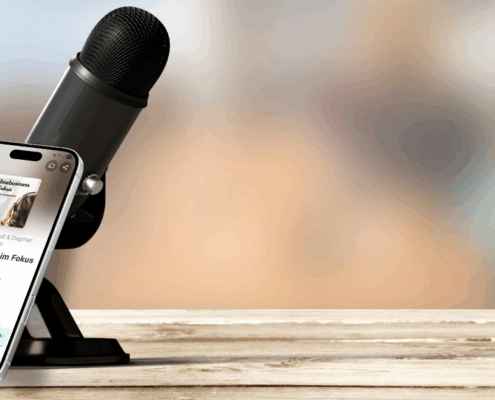 Ein Mikrofon auf einem Ständer steht auf einer Holzfläche neben einem Smartphone, auf dem der Podcast "Onlinebusiness im Fokus" angezeigt wird. Die Szene fängt modernes Hörverhalten ein, in der rechten unteren Ecke steht "MICRO AUDIO SYMPOSIUM".