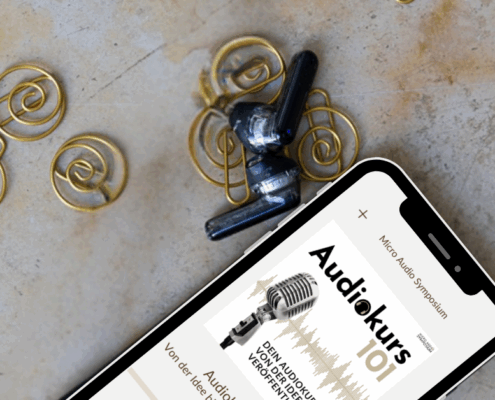 Ein Smartphone, das einen "Audiokurs 101" mit dem Bild eines alten Mikrofons anzeigt, liegt auf einer Marmorfläche neben spiralförmigen goldenen Drahtohrringen und schwarzen kabellosen Kopfhörern. In der Ecke ist "MICRO AUDIO SYMPOSIUM" aufgedruckt.