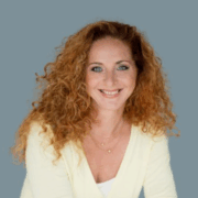 MAS Silvia Chytil Eine Frau mit langen, lockigen roten Haaren und blauen Augen lächelt in die Kamera. Sie trägt einen hellgelben Pullover über einem weißen Oberteil, mehrere Ketten und ein diskretes Micro Audio-Gerät vor einem schlichten blaugrauen Hintergrund.