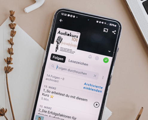 Ein Smartphone, auf dessen Bildschirm eine Podcast-App mit "Audiekurs 101" und seinem Glossar angezeigt wird, liegt auf einer hellen Fläche neben einer Laptop-Tastatur, einer getrockneten Pflanze und einer weißen Notizkarte. Auf dem Cover des Podcasts ist ein Mikrofon abgebildet.