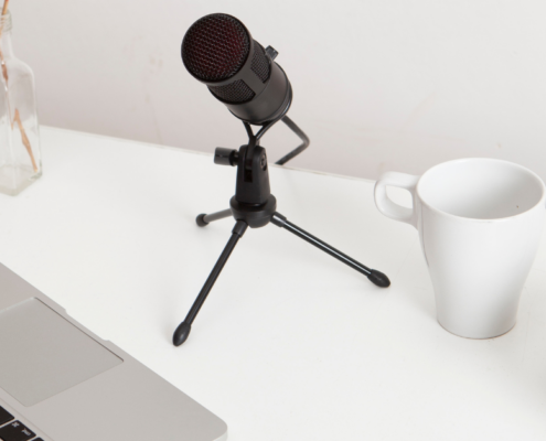 Ein schwarzes Mikrofon auf einem Stativ steht auf einem weißen Schreibtisch neben einem Laptop, einer weißen Tasse, Kopfhörern und einer Glasvase mit getrockneten Zweigen - perfekt für Podcast-Marketing-Sessions. In der unteren rechten Ecke erscheint der Text "MICRO AUDIO SYMPOSIUM".