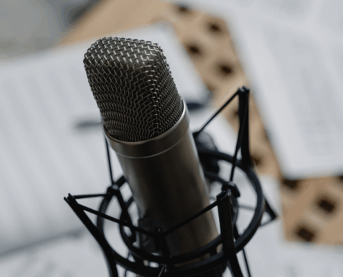 Eine Nahaufnahme eines professionellen Mikrofons auf einem Ständer, mit unscharfen Papierblättern im Hintergrund. In der unteren rechten Ecke ist "MICRO AUDIO SYMPOSIUM" zu sehen, was die Atmosphäre einer Podcast-Aufnahme heraufbeschwört.