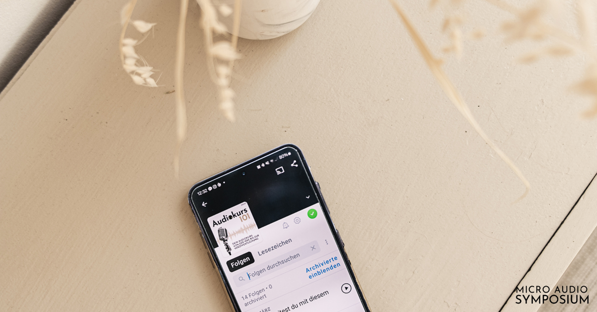 Ein Smartphone mit einer Podcast-App liegt auf einem hellen Tisch neben einer weißen Vase mit getrockneten beigen Pflanzen und spiegelt die Prinzipien des minimalistischen Designs wider. In der rechten unteren Ecke steht der Schriftzug "MICRO AUDIO SYMPOSIUM".