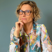 MAS Sandra Holze Eine Frau mit kurzem, lockigem, blondem Haar, einer großen schwarzen Brille und einem bunt gemusterten Hemd sitzt vor einem schlichten Hintergrund, stützt ihr Kinn auf die Hand und lächelt nachdenklich.
