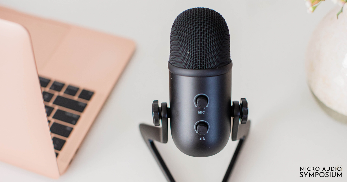 Ein schwarzes Mikrofon auf einem Ständer steht auf einem weißen Schreibtisch neben einem roségoldenen Laptop, was darauf hindeutet, dass das Hörverhalten im Mittelpunkt steht. In der rechten unteren Ecke steht der Schriftzug "MICRO AUDIO SYMPOSIUM".