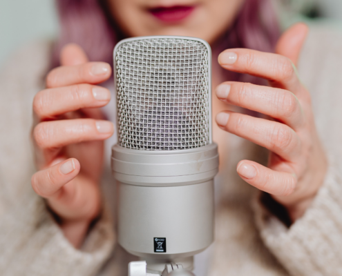 Eine Person mit hellrosa Nägeln hält sanft ein großes silbernes Kondensatormikrofon, das auf Podcasting 2025 hinweist, mit "MICRO AUDIO SYMPOSIUM" in der unteren rechten Ecke. Der sanft verschwommene Hintergrund verleiht dem Bild einen modernen Touch.