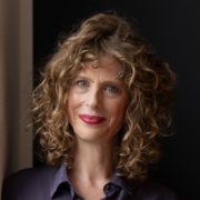 MAS Miriam Betancourt Eine Frau mit lockigem, schulterlangem hellbraunem Haar und blauen Augen lächelt sanft. Sie trägt eine violette Bluse und rosa Lippenstift und steht in weichem Licht vor einem dunklen Hintergrund.