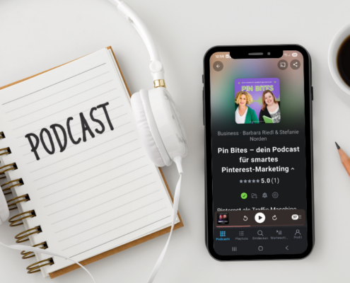 Ein Smartphone, das eine Podcast-Episode mit einem leuchtenden Podcast-Cover anzeigt, liegt auf einem weißen Schreibtisch neben einem Spiralnotizbuch mit der Aufschrift "PODCAST" und weißen Kopfhörern. In der Ecke ist das Logo des "Micro Audio Symposium" zu sehen.