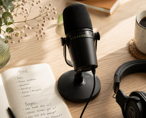 Ein privates Podcast-Setup auf einem Holztisch mit Mikrofon, Kopfhörern, einem Notizbuch mit handschriftlichen Notizen, einer Tasse Kaffee auf einem Untersetzer und einer Vase mit kleinen Blumen. Sonnenlicht fällt auf den Arbeitsbereich.