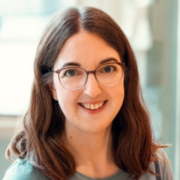 Eine Frau mit langen braunen Haaren und Brille lächelt in die Kamera. Sie trägt ein helles Oberteil und scheint in einem Innenraum mit weichem, unscharfem Hintergrund ein Micro Audio Gerät zu benutzen.