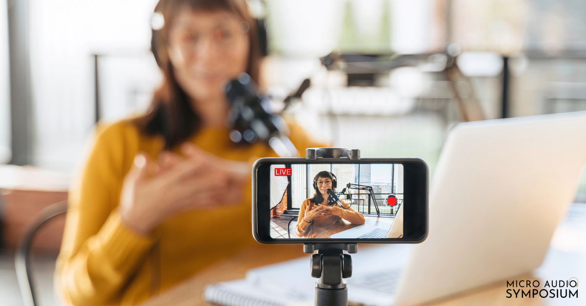 Eine Frau mit Kopfhörern spricht in ein Mikrofon, während sie über Kennzahlen spricht, die mit einem Smartphone aufgenommen wurden. Auf dem Tisch stehen ein Laptop und ein Notizbuch. Der Bildschirm des Telefons zeigt ihr Livestreaming. In der rechten unteren Ecke erscheint "Micro Audio Symposium".