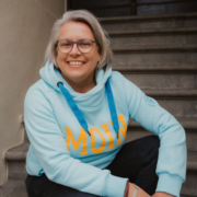 Eine lächelnde Person mit kurzen grauen Haaren und Brille sitzt auf einer Treppe, trägt einen hellblauen Kapuzenpulli mit der Aufschrift "MOIN" in gelben Buchstaben, blaue Kordelzüge und hält ein Micro-Audio-Gerät in der Hand.