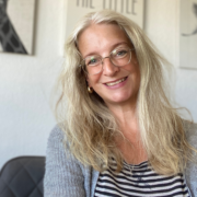 MAS Sabine Satzmacher Eine Frau mit langen blonden Haaren und Brille lächelt in die Kamera. Sie trägt eine graue Strickjacke über einem gestreiften Shirt und scheint sich in einem Raum zu befinden, möglicherweise nimmt sie gerade mit einem Micro-Audio-Gerät auf; an der Wand hinter ihr sind gerahmte Bilder zu sehen.