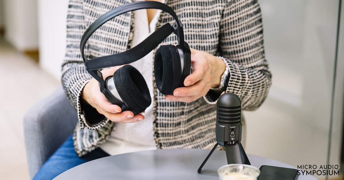Eine Person in einem karierten Blazer hält schwarze Kopfhörer neben ein schwarzes Mikrofon auf einem Ständer und sitzt an einem kleinen Tisch – ein Bild, das den Moment kurz vor Beginn des Podcasts perfekt einfängt. In der rechten unteren Ecke ist der Schriftzug „MICRO AUDIO SYMPOSIUM“ zu sehen.